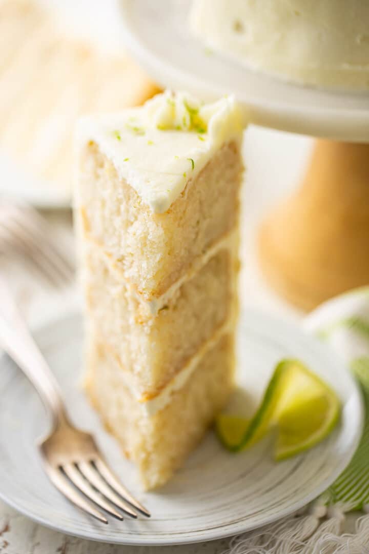 Keylime cake recipe, baked, frosted, and cut into a triangular slice, garnished with a slice of fresh lime.