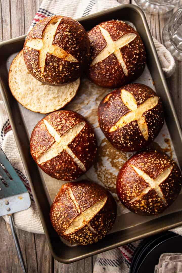 Overhead image of pretzel bun recipe, prepared, baked and presented on a parchment-lined baking sheet with beer glasses and a grill spatula.