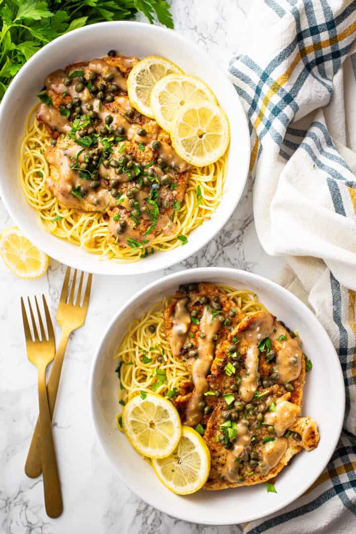 Two bowls of lemon chicken piccata on a white marble surface.