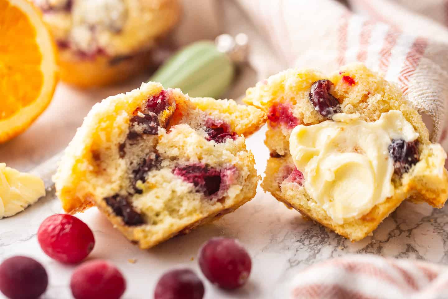 Cranberry orange muffin buttered, with a fresh orange in the background and a red striped kitchen towel.