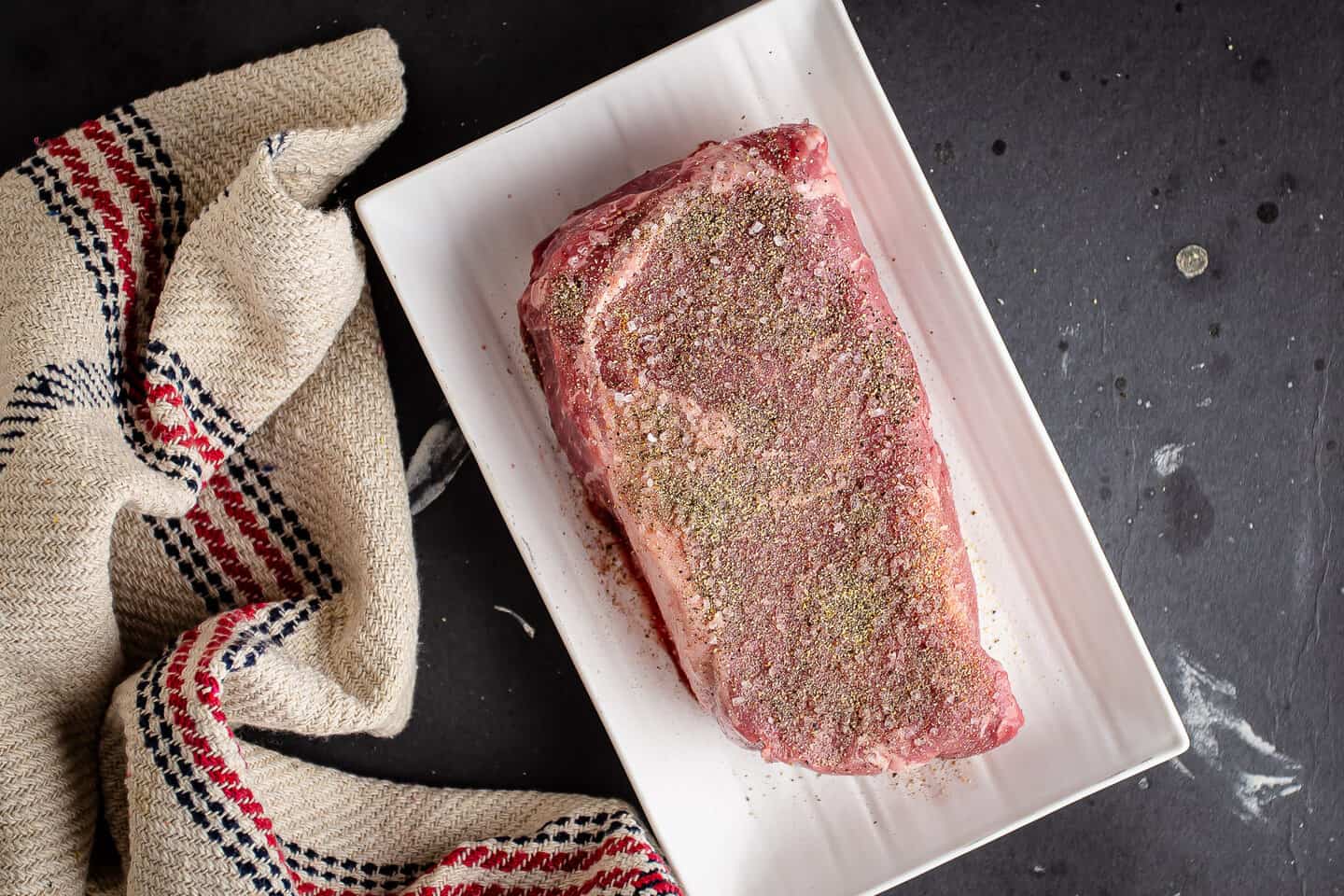 A generously seasoned ribeye steak on a white tray.
