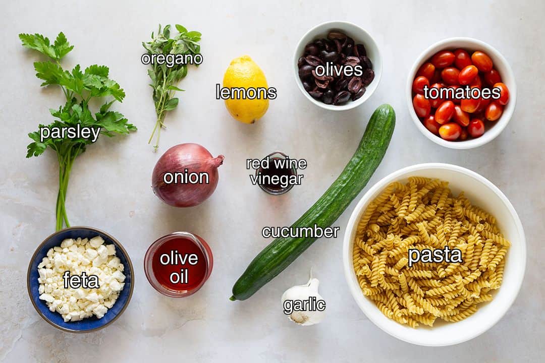 Ingredients with text overlay for Greek orzo pasta salad, including parsley, oregano, lemons, olives, tomatoes, onion, vinegar, feta, olive oil, cucumber, garlic, and pasta.