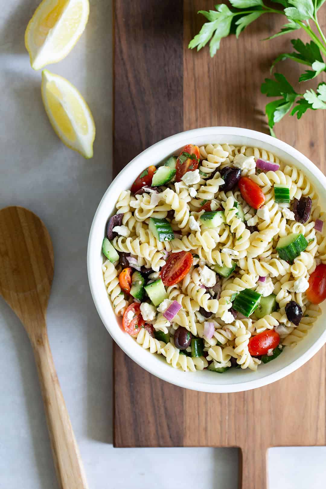 Recipe for Greek pasta salad in a bowl set on a cutting board.