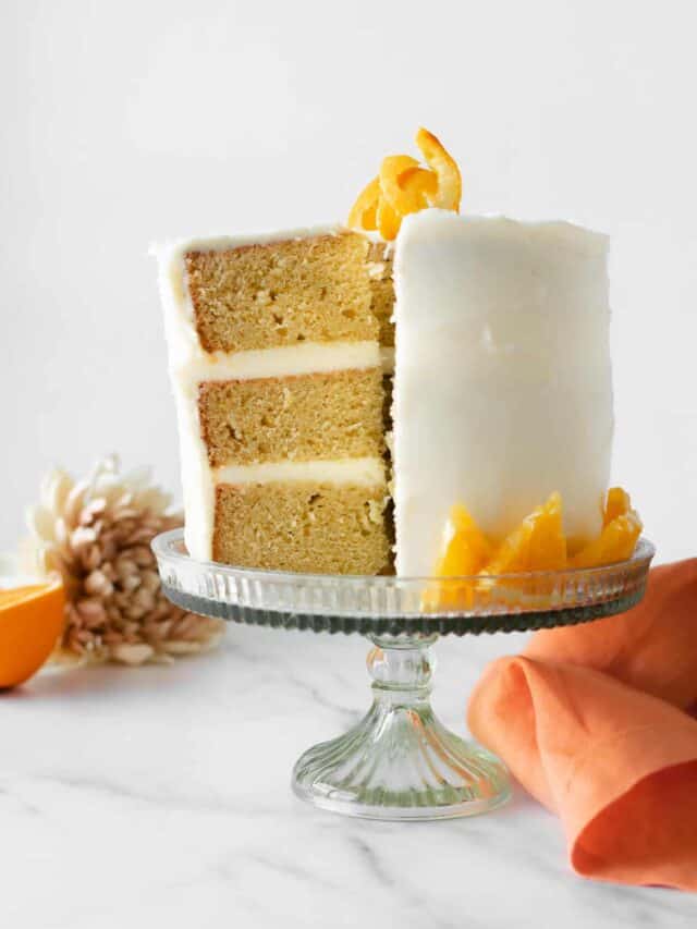 three layer orange cake with cream cheese frosting on a cake stand with an orange napkin.