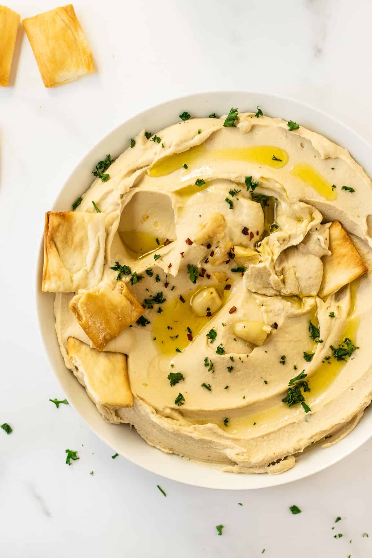hummus recipe in a white bowl topped with parsley, olive oil and pita chips.