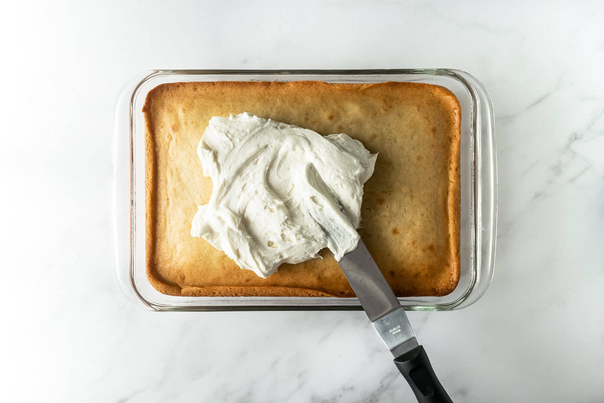 vanilla cake in a baking pan with vanilla frosting and an offset spatula.