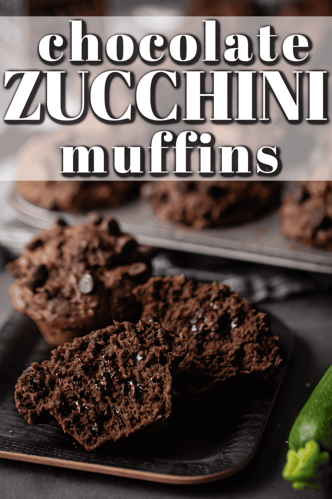 Two chocolate zucchini muffins on a dark plate, one of them split in half to display the melty chocolate chips.