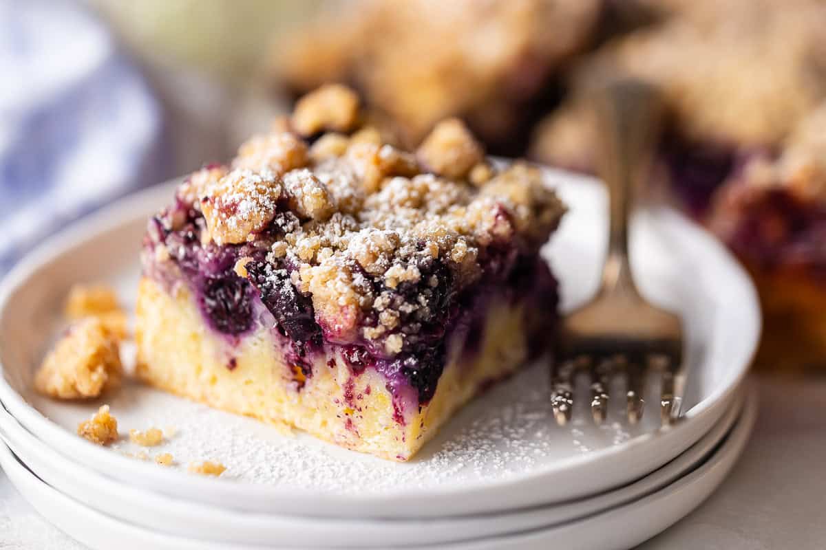 A slice of blueberry buckle, topped with powdered sugar and presented on a white plate with a vintage silver fork.