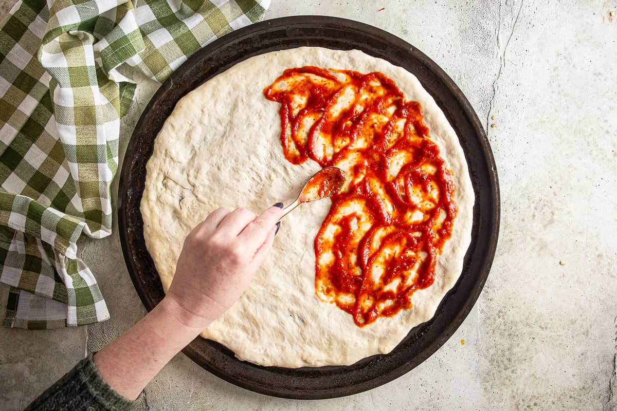Adding a thin layer of pizza sauce onto stretched dough for jalapeno popper pizza.