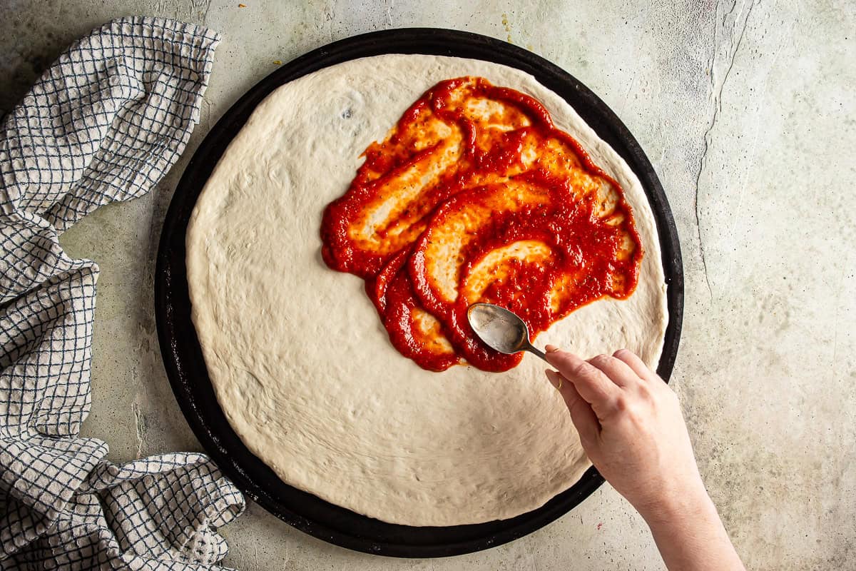 Sauce being spread on stretched pizza dough.