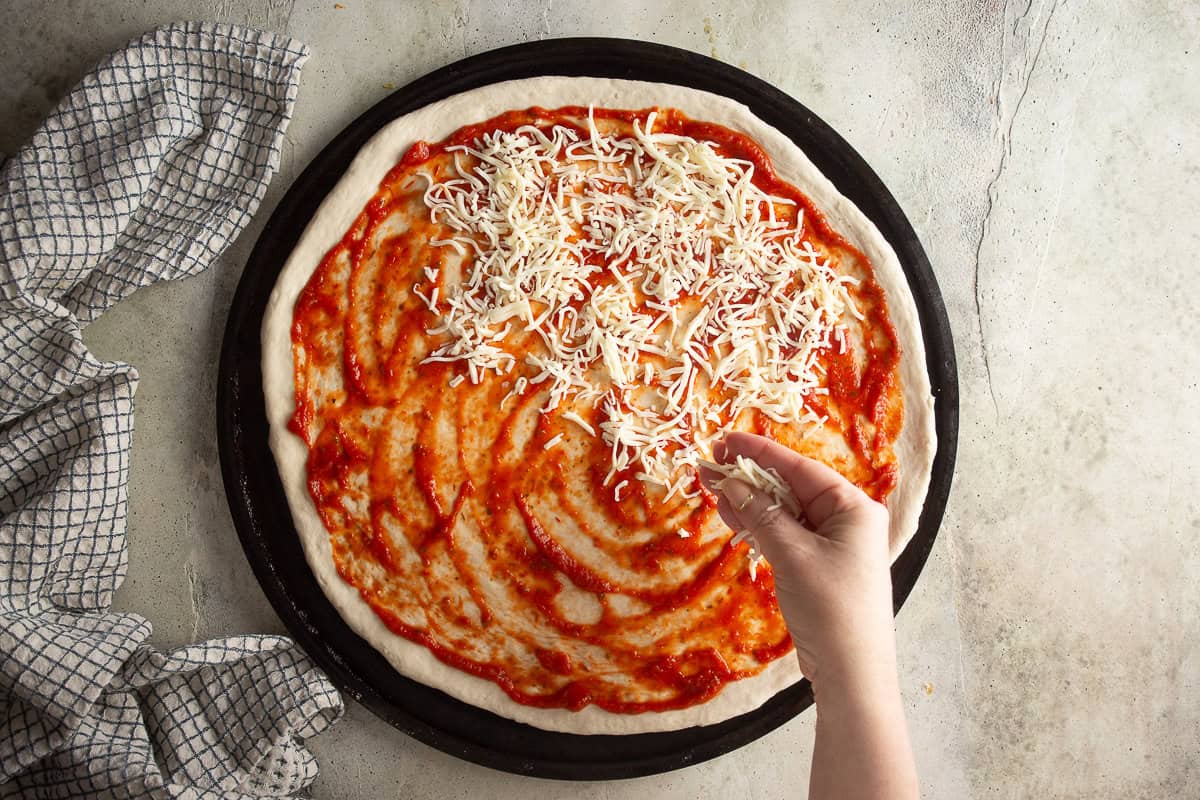 Mozzarella cheese being sprinkled over a pizza crust topped with sauce.
