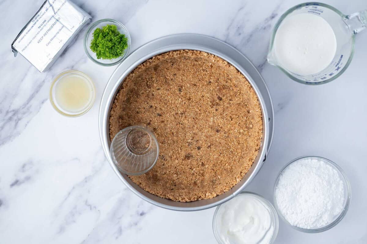 graham cracker crust being pressed into a springform pan with a cup