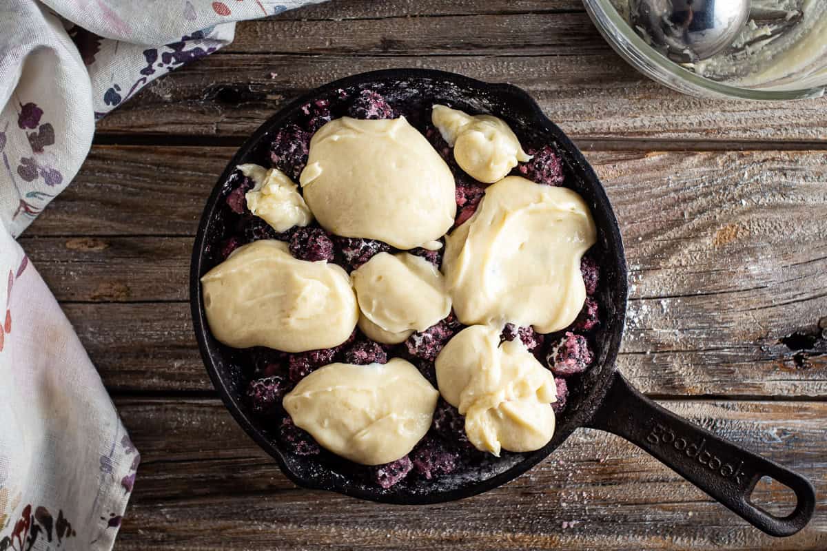 Adding cobbler batter on top of the coated blackberries in a cast iron skillet.