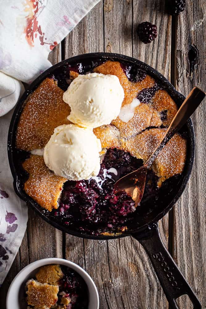Blackberry cobbler in a cast iron skillet with ice cream scooped on top and a spoon in the dish.