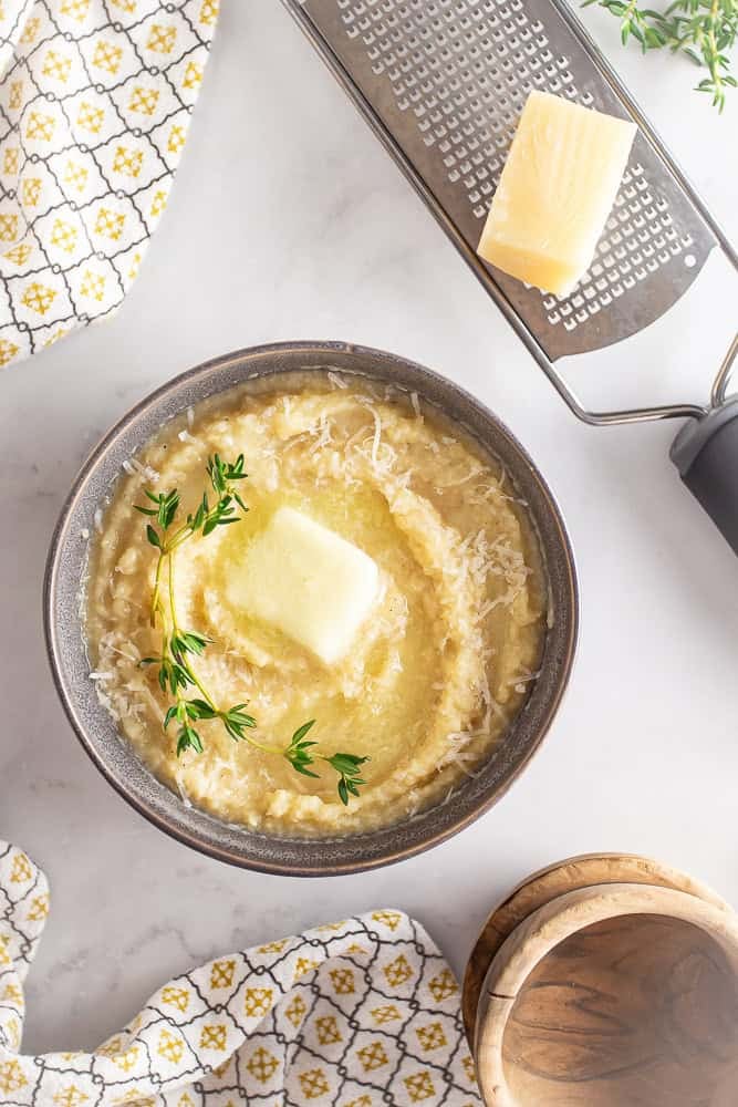 Polenta in a serving dish with butter and thyme and a cheese grater with parmesan near the bowl.
