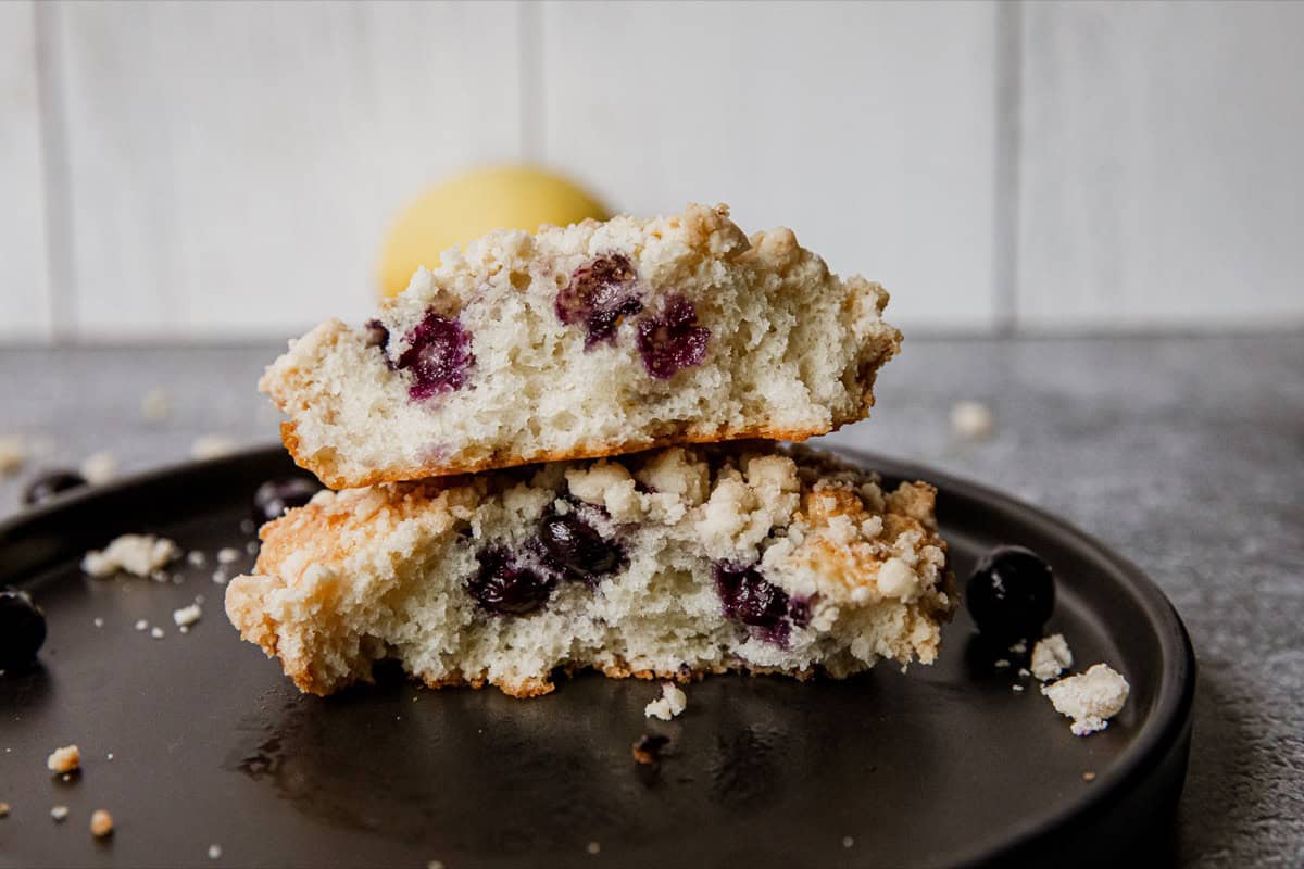 Close-up of blueberry streusel muffin tops on black plate with crumbs and blueberries.
