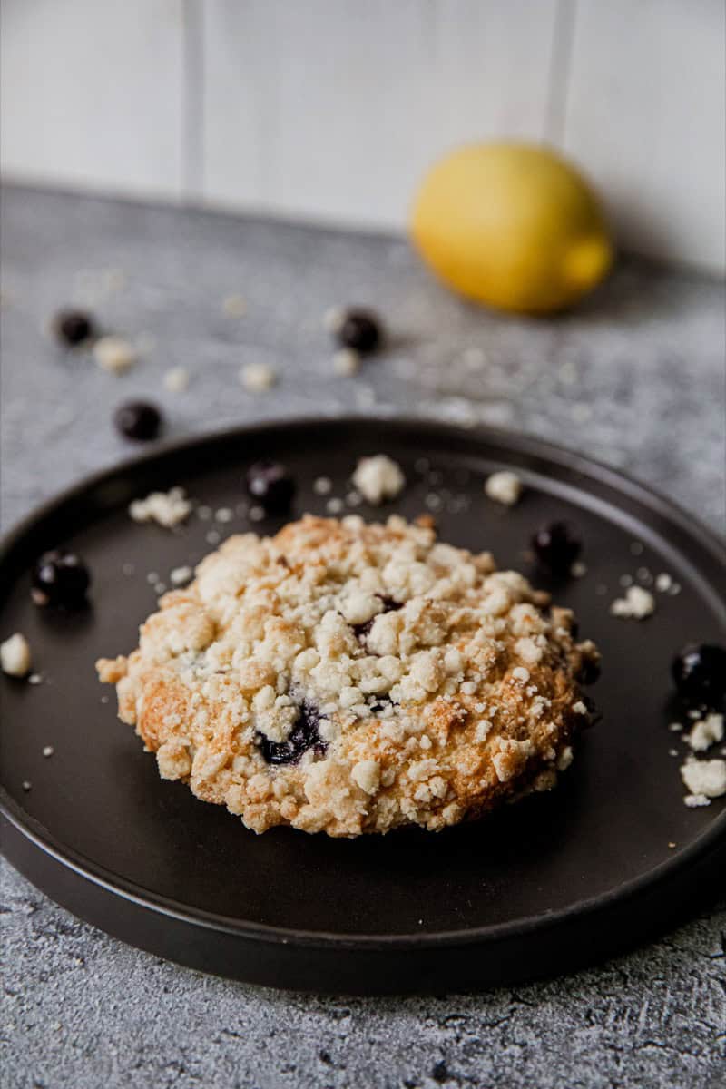 Close-up of a blueberry streusel muffin top on a black plate with lemon in background.