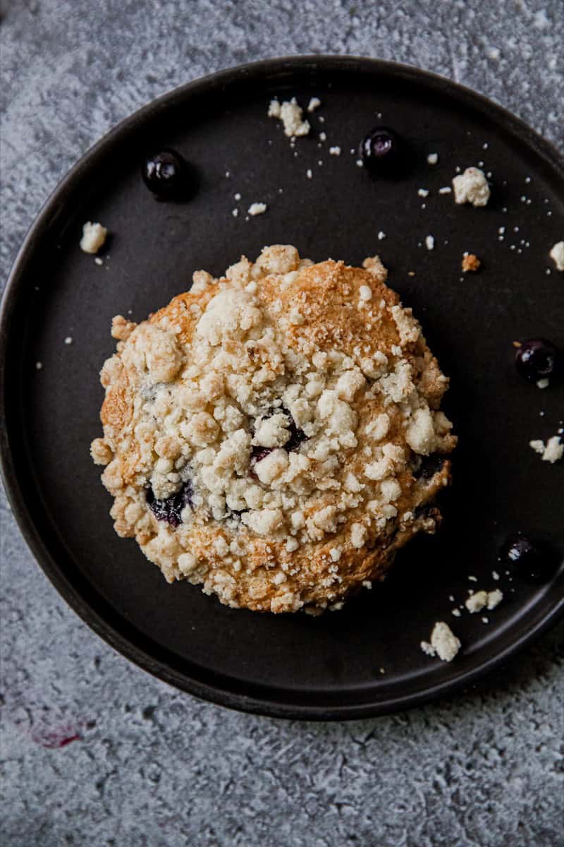 Blueberry streusel muffin top on black plate with blueberries around it.