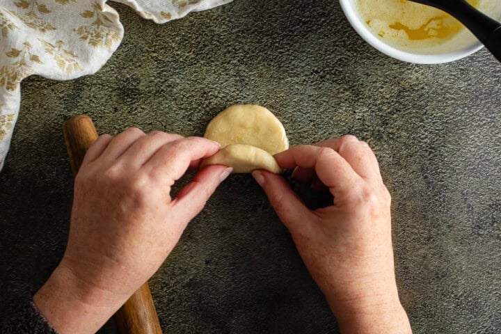 Hands shaping dough for Parker House Rolls on a textured surface with ingredients nearby.