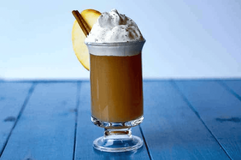 Caramel apple cocktail with whipped cream and cinnamon stick on blue table.
