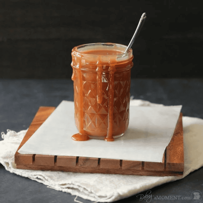 Creamy caramel sauce in a glass jar with a spoon on a wooden board.