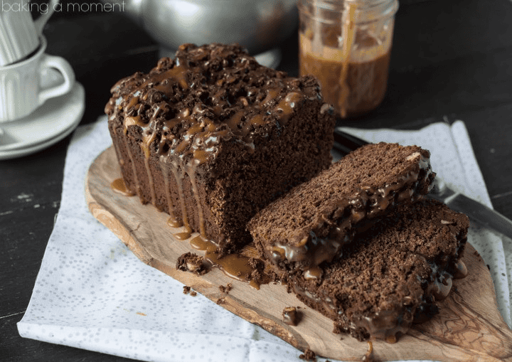 Chocolate cake with caramel drizzle and chocolate chips on top, sliced on a wooden board.