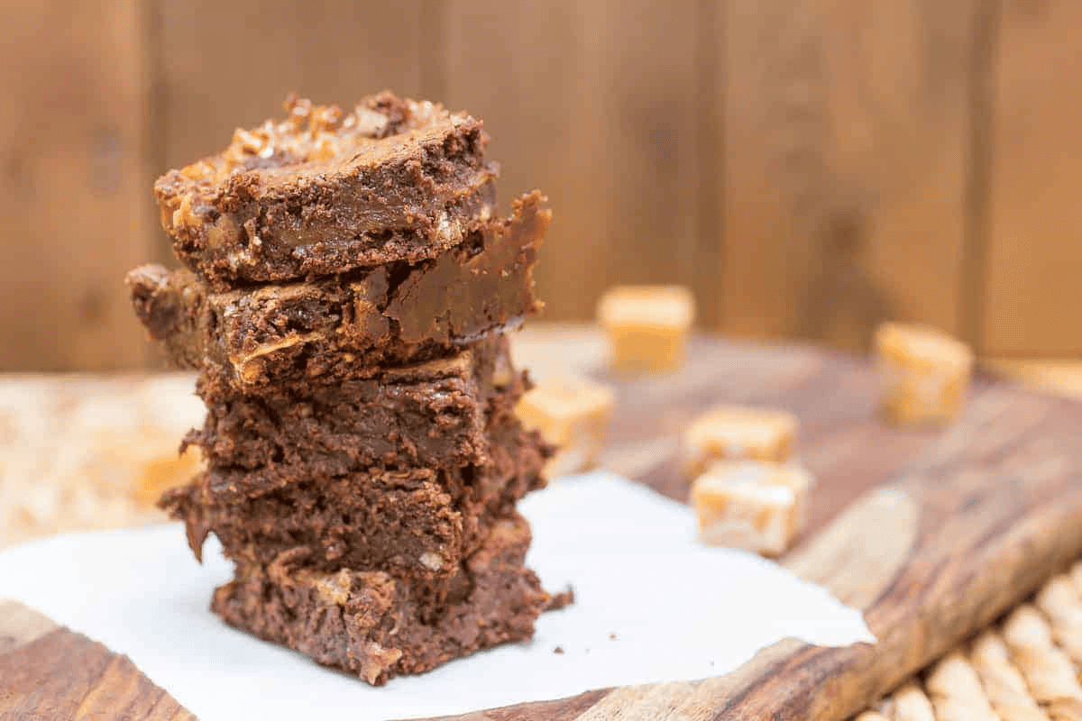 Stacked caramel brownies on a wooden board with caramel pieces in the background.